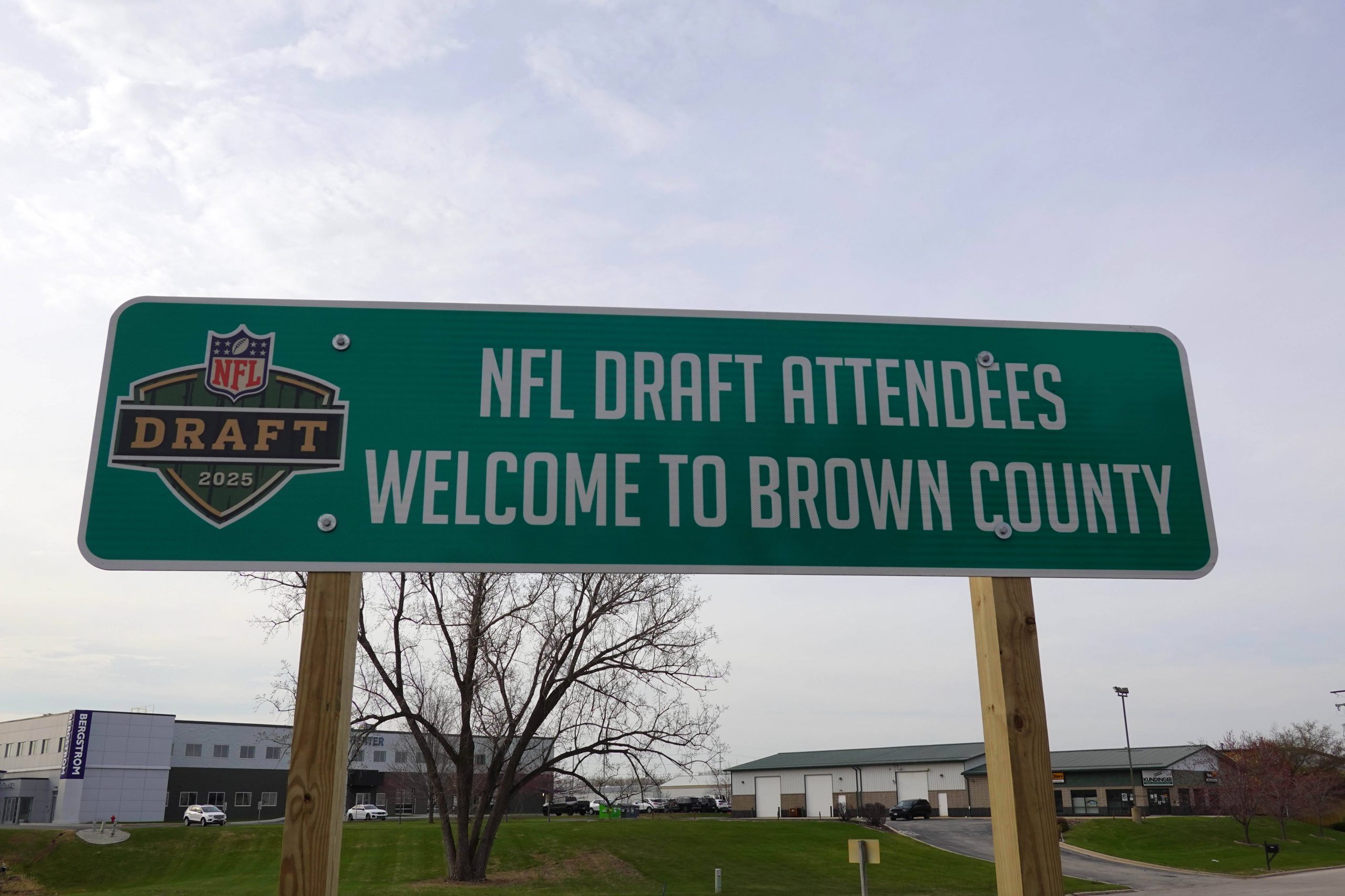 NFL Draft 2025 - Cleveland Browns und New York Giants planen Trades. Auf einem grünen Schild am Straßenrand mit dem Logo des NFL Draft 2025 steht „Willkommen in Brown County, Teilnehmer des NFL Draft“. Das Schild ist auf Holzpfosten montiert und steht vor einer Kulisse aus Gebäuden, Gras und einem kahlen Baum. Diese Beschreibung wurde mit der FootballR KI automatisch generiert.