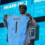 Cam Ward in Anzug und blaugrünem Hut hält beim NFL Draft 2025 auf der Bühne ein hellblaues Footballtrikot mit dem Namen „WARD“ und der Nummer 1 hoch, während Cam Ward auf einer hellen Leinwand dahinter gefeiert wird, auf der „MIAMI“ und Grafiken zum Thema Draft zu sehen sind. Diese Beschreibung wurde mit der FootballR KI automatisch generiert.