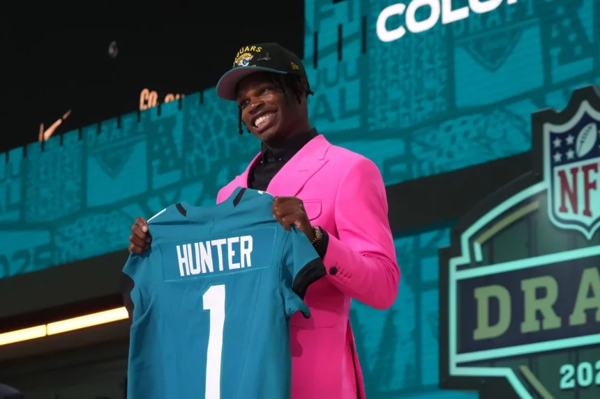 Ein lächelnder junger Mann, Travis Hunter, in einem leuchtend rosa Anzug und einer Teamkappe, hält ein blaugrünes Footballtrikot mit „HUNTER“ und der Nummer 1 hoch. Hinter ihm befindet sich eine NFL Draft-Bühne mit Logos und blaugrünen Grafiken, die seine Draft-Auswahl kennzeichnen. Diese Beschreibung wurde mit der FootballR KI automatisch generiert.