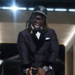 Ashton Jeanty in einem dunkel gemusterten Smoking, mit Fliege und Brille lacht fröhlich. Er trägt eine schwarze Raiders-Kappe und steht vor einem leuchtend gelben Gebäude. Mit gefalteten Händen und wehenden langen Dreadlocks feiert er den NFL Draft 2025. Diese Beschreibung wurde mit der FootballR KI automatisch generiert.