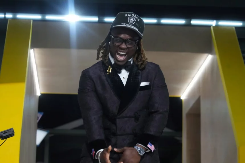 Ashton Jeanty in einem dunkel gemusterten Smoking, mit Fliege und Brille lacht fröhlich. Er trägt eine schwarze Raiders-Kappe und steht vor einem leuchtend gelben Gebäude. Mit gefalteten Händen und wehenden langen Dreadlocks feiert er den NFL Draft 2025. Diese Beschreibung wurde mit der FootballR KI automatisch generiert.