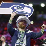 Im Free TV - Ein Fan in Trikot und Mütze der Seattle Seahawks jubelt beim NFL Draft 2025 in der Menge und hält ein großes Schaumstoffschild in Form des Seahawks-Logos hoch. Er trägt Perlenketten und Armbänder, im Hintergrund sind bunte Lichter und andere Fans zu sehen. Diese Beschreibung wurde mit der FootballR KI automatisch generiert.