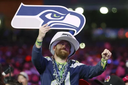 Im Free TV - Ein Fan in Trikot und Mütze der Seattle Seahawks jubelt beim NFL Draft 2025 in der Menge und hält ein großes Schaumstoffschild in Form des Seahawks-Logos hoch. Er trägt Perlenketten und Armbänder, im Hintergrund sind bunte Lichter und andere Fans zu sehen. Diese Beschreibung wurde mit der FootballR KI automatisch generiert.