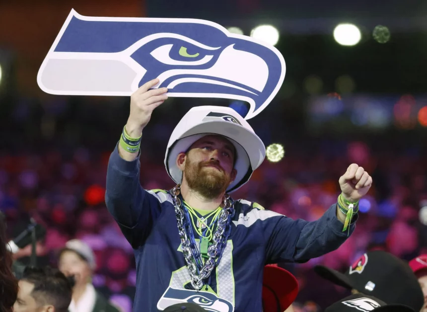 Im Free TV - Ein Fan in Trikot und Mütze der Seattle Seahawks jubelt beim NFL Draft 2025 in der Menge und hält ein großes Schaumstoffschild in Form des Seahawks-Logos hoch. Er trägt Perlenketten und Armbänder, im Hintergrund sind bunte Lichter und andere Fans zu sehen. Diese Beschreibung wurde mit der FootballR KI automatisch generiert.