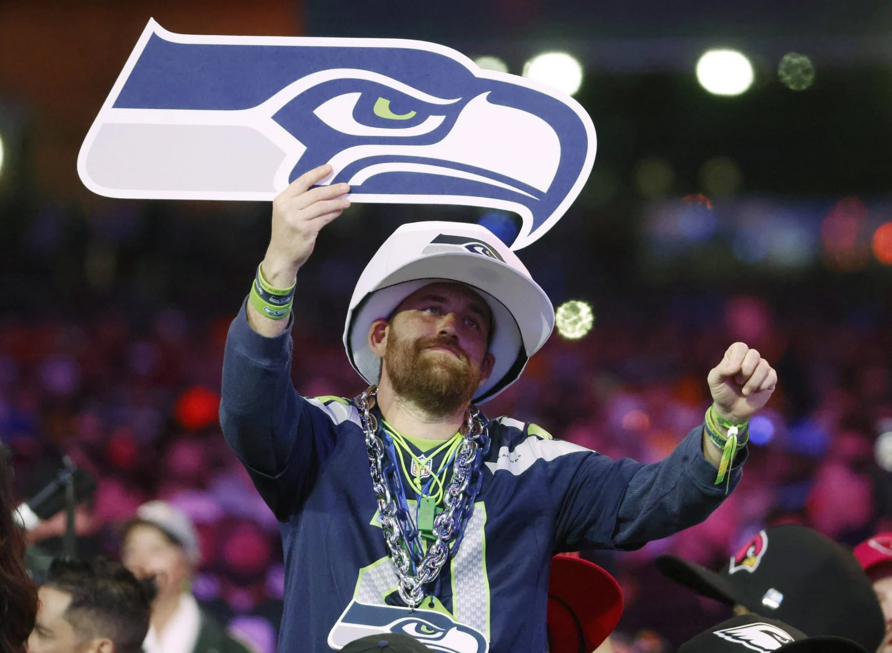 Im Free TV - Ein Fan in Trikot und Mütze der Seattle Seahawks jubelt beim NFL Draft 2025 in der Menge und hält ein großes Schaumstoffschild in Form des Seahawks-Logos hoch. Er trägt Perlenketten und Armbänder, im Hintergrund sind bunte Lichter und andere Fans zu sehen. Diese Beschreibung wurde mit der FootballR KI automatisch generiert.