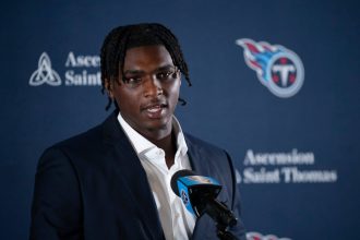 Cam Ward mit kurzen Dreadlocks, einem dunklen Anzug und einem weißen Hemd, spricht in ein Mikrofon mit dem Logo der Tennessee Titans. Im Hintergrund sind das Titans-Logo und die Schriftzüge „Ascension Saint Thomas“ auf blauem Grund zu sehen. Diese Beschreibung wurde mit der FootballR KI automatisch generiert.