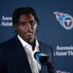 Cam Ward mit kurzen Dreadlocks, einem dunklen Anzug und einem weißen Hemd, spricht in ein Mikrofon mit dem Logo der Tennessee Titans. Im Hintergrund sind das Titans-Logo und die Schriftzüge „Ascension Saint Thomas“ auf blauem Grund zu sehen. Diese Beschreibung wurde mit der FootballR KI automatisch generiert.