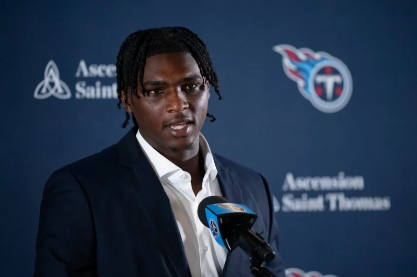 Cam Ward mit kurzen Dreadlocks, einem dunklen Anzug und einem weißen Hemd, spricht in ein Mikrofon mit dem Logo der Tennessee Titans. Im Hintergrund sind das Titans-Logo und die Schriftzüge „Ascension Saint Thomas“ auf blauem Grund zu sehen. Diese Beschreibung wurde mit der FootballR KI automatisch generiert.