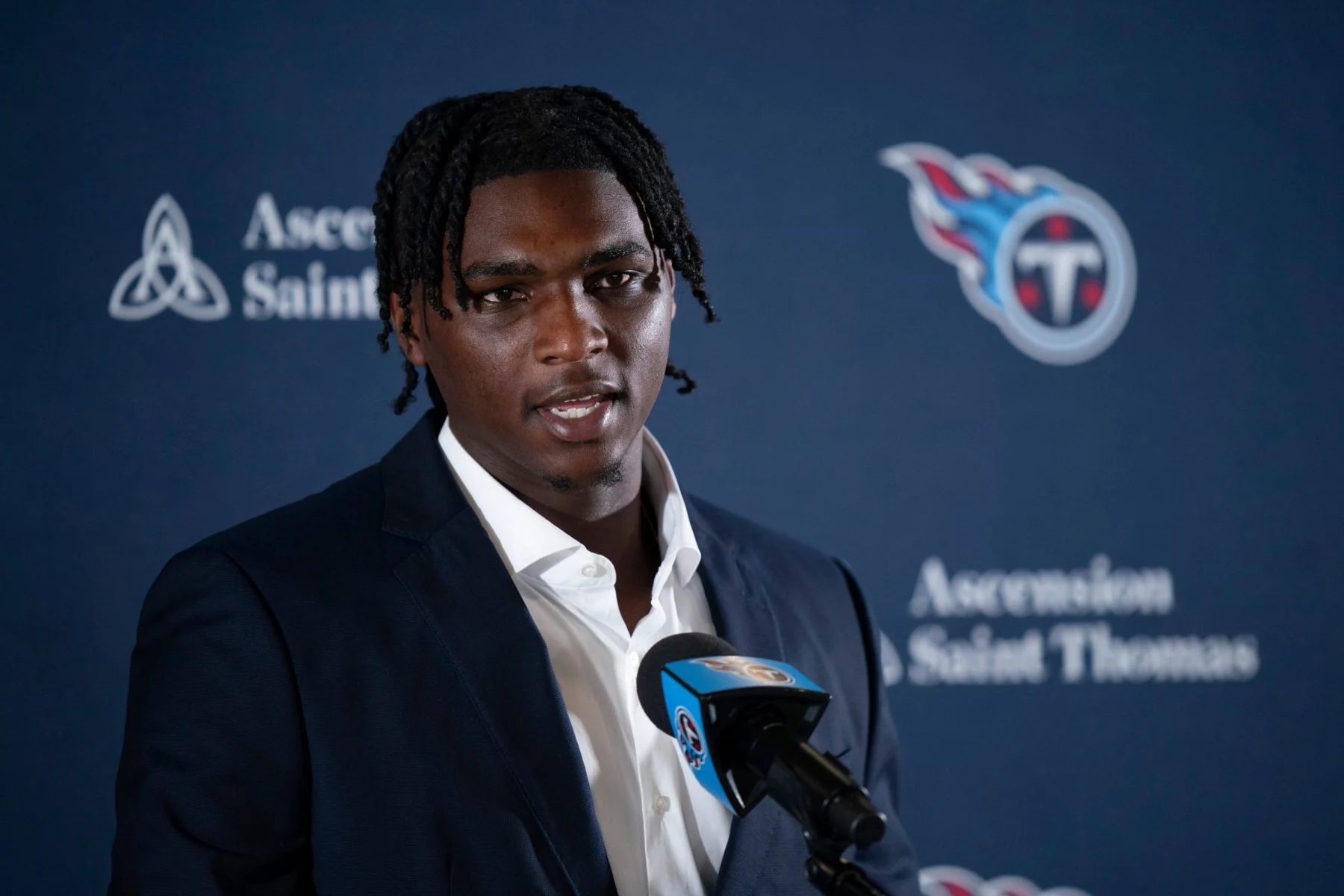 Cam Ward mit kurzen Dreadlocks, einem dunklen Anzug und einem weißen Hemd, spricht in ein Mikrofon mit dem Logo der Tennessee Titans. Im Hintergrund sind das Titans-Logo und die Schriftzüge „Ascension Saint Thomas“ auf blauem Grund zu sehen. Diese Beschreibung wurde mit der FootballR KI automatisch generiert.