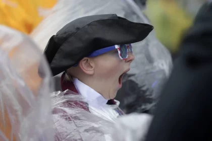 Ein Kind mit Dreispitz, Kolonialkleidung und patriotischer Sonnenbrille gähnt herzhaft. Kobee Minor ist teilweise von einem durchsichtigen Plastikregenponcho bedeckt. Im Hintergrund sind Menschen und Gelb unscharf zu sehen, was auf eine verregnete Veranstaltung im Freien hindeutet. Diese Beschreibung wurde mit der FootballR KI automatisch generiert.