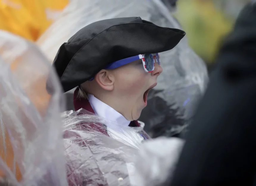 Ein Kind mit Dreispitz, Kolonialkleidung und patriotischer Sonnenbrille gähnt herzhaft. Kobee Minor ist teilweise von einem durchsichtigen Plastikregenponcho bedeckt. Im Hintergrund sind Menschen und Gelb unscharf zu sehen, was auf eine verregnete Veranstaltung im Freien hindeutet. Diese Beschreibung wurde mit der FootballR KI automatisch generiert.