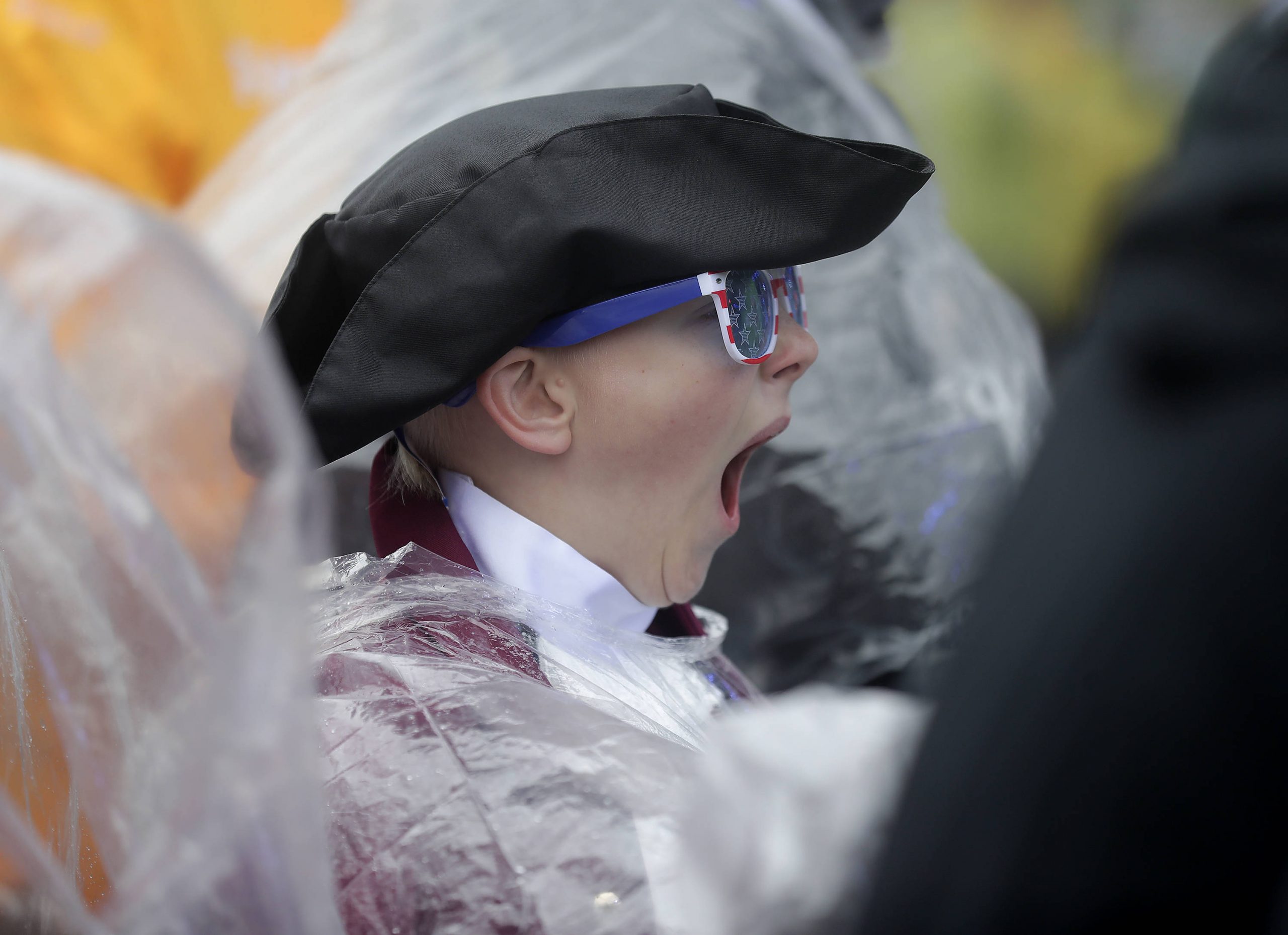 Ein Kind mit Dreispitz, Kolonialkleidung und patriotischer Sonnenbrille gähnt herzhaft. Kobee Minor ist teilweise von einem durchsichtigen Plastikregenponcho bedeckt. Im Hintergrund sind Menschen und Gelb unscharf zu sehen, was auf eine verregnete Veranstaltung im Freien hindeutet. Diese Beschreibung wurde mit der FootballR KI automatisch generiert.
