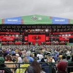 Shedeur Sanders - Eine große Menschenmenge in Regenponchos steht vor einer großen, hell erleuchteten Bühne für den NFL Draft 2021. Auf der Bühne hängen grüne Banner mit den Aufschriften „BUD LIGHT“ und „NFL DRAFT 2021“, während rote digitale Bildschirme überraschend verkünden: „BROWNS ON THE CLOCK“. Darüber ragt ein bedeckter Himmel empor. Diese Beschreibung wurde mit der FootballR KI automatisch generiert.