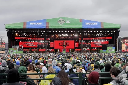 Shedeur Sanders - Eine große Menschenmenge in Regenponchos steht vor einer großen, hell erleuchteten Bühne für den NFL Draft 2021. Auf der Bühne hängen grüne Banner mit den Aufschriften „BUD LIGHT“ und „NFL DRAFT 2021“, während rote digitale Bildschirme überraschend verkünden: „BROWNS ON THE CLOCK“. Darüber ragt ein bedeckter Himmel empor. Diese Beschreibung wurde mit der FootballR KI automatisch generiert.