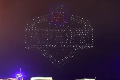 Derrick Harmon - Ein Nachthimmel wird von Hunderten Drohnen erleuchtet, die das NFL Draft-Logo bilden. „DRAFT“, das NFL-Schild und Derrick Harmons Name sind in Lichtern über grün dekorierten Gebäuden und dem „G“-Logo der Packers zu sehen. Diese Beschreibung wurde mit der FootballR KI automatisch generiert.