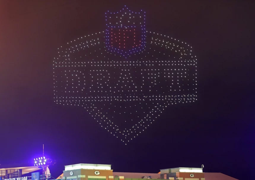 Derrick Harmon - Ein Nachthimmel wird von Hunderten Drohnen erleuchtet, die das NFL Draft-Logo bilden. „DRAFT“, das NFL-Schild und Derrick Harmons Name sind in Lichtern über grün dekorierten Gebäuden und dem „G“-Logo der Packers zu sehen. Diese Beschreibung wurde mit der FootballR KI automatisch generiert.