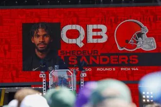 100 Millionen - Auf einem großen Bildschirm ist „QB Shedeur Sanders“ mit seinem Porträt und einem orangefarbenen Footballhelm zu sehen. Darunter steht „Runde 5, Pick 144“. Shedeur Sanders ist bei einem Event, das wie ein NFL-Draft-Event aussieht, prominent vertreten, umgeben von Menschen und Ausrüstung. Diese Beschreibung wurde mit der FootballR KI automatisch generiert.