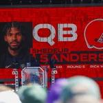 100 Millionen - Auf einem großen Bildschirm ist „QB Shedeur Sanders“ mit seinem Porträt und einem orangefarbenen Footballhelm zu sehen. Darunter steht „Runde 5, Pick 144“. Shedeur Sanders ist bei einem Event, das wie ein NFL-Draft-Event aussieht, prominent vertreten, umgeben von Menschen und Ausrüstung. Diese Beschreibung wurde mit der FootballR KI automatisch generiert.