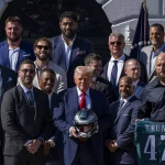 Eagles im Weißen Haus - Eine Gruppe von Männern in Anzügen und Hemden mit Kragen steht draußen in der Nähe des Weißen Hauses. Ein Mann in der Mitte hält einen American-Football-Helm, ein anderer trägt ein dunkelgrünes „TRUMP 47“-Eagles-Trikot und feiert den 19. Super Bowl. Im Hintergrund wehen amerikanische Flaggen. Diese Beschreibung wurde mit der FootballR KI automatisch generiert.