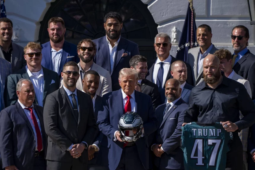 Eagles im Weißen Haus - Eine Gruppe von Männern in Anzügen und Hemden mit Kragen steht draußen in der Nähe des Weißen Hauses. Ein Mann in der Mitte hält einen American-Football-Helm, ein anderer trägt ein dunkelgrünes „TRUMP 47“-Eagles-Trikot und feiert den 19. Super Bowl. Im Hintergrund wehen amerikanische Flaggen. Diese Beschreibung wurde mit der FootballR KI automatisch generiert.