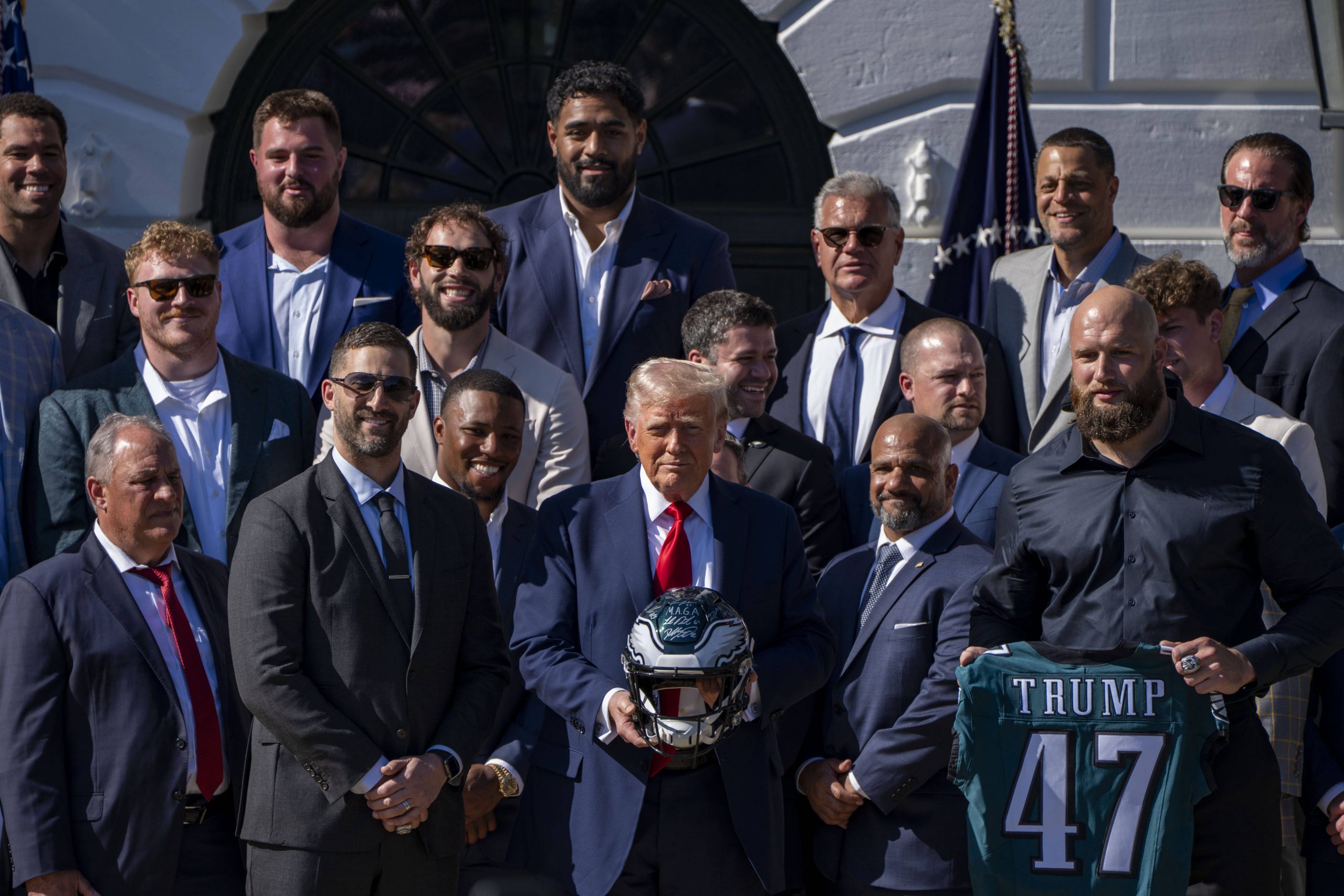 Eagles im Weißen Haus - Eine Gruppe von Männern in Anzügen und Hemden mit Kragen steht draußen in der Nähe des Weißen Hauses. Ein Mann in der Mitte hält einen American-Football-Helm, ein anderer trägt ein dunkelgrünes „TRUMP 47“-Eagles-Trikot und feiert den 19. Super Bowl. Im Hintergrund wehen amerikanische Flaggen. Diese Beschreibung wurde mit der FootballR KI automatisch generiert.