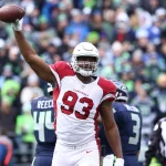 Calais Campbell im rot-weißen Trikot der Arizona Cardinals mit der Nummer 93 reckt während eines Spiels triumphierend einen Football in die Höhe. Es ist niemand Geringeres als Calais Campbell. Im Hintergrund sind verschwommen Spieler der Seattle Seahawks und ein Schiedsrichter zu sehen, zusammen mit einer Menge Fans. Diese Beschreibung wurde mit der FootballR KI automatisch generiert.