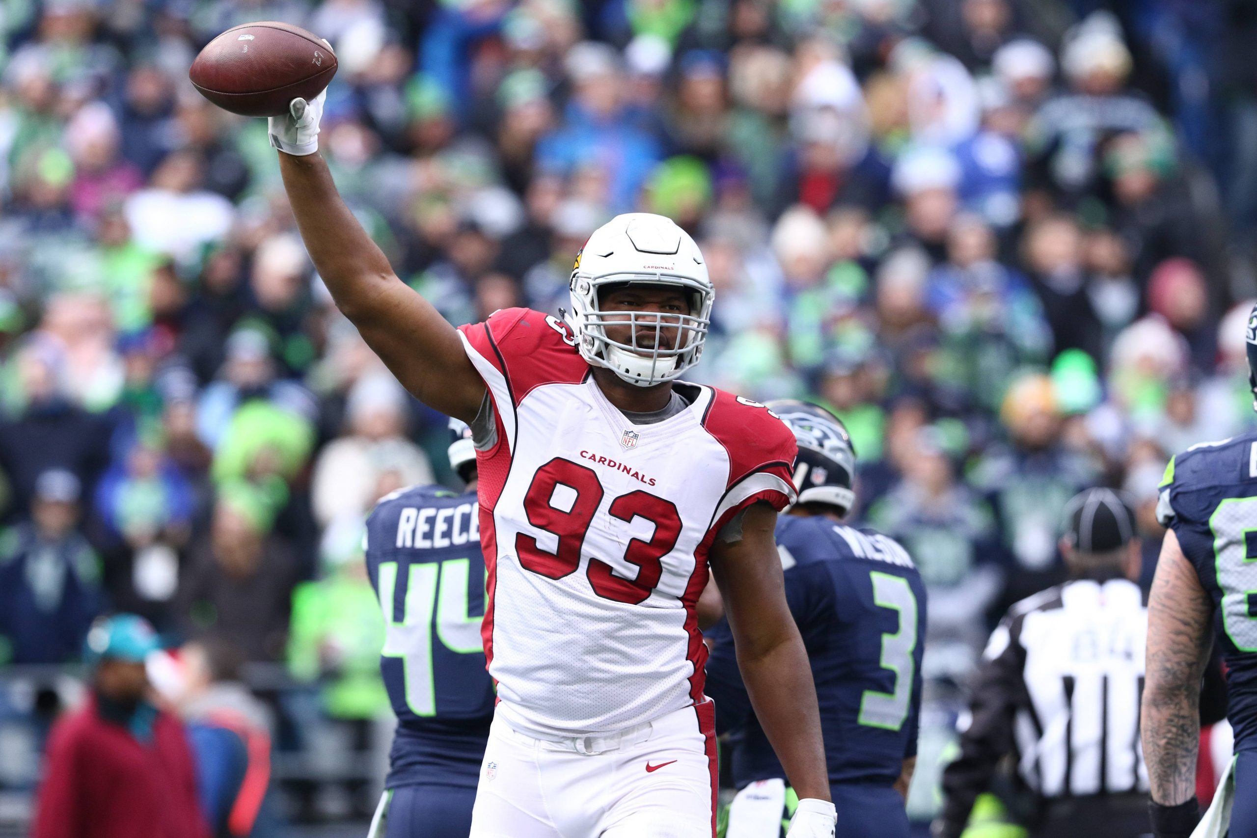 Calais Campbell im rot-weißen Trikot der Arizona Cardinals mit der Nummer 93 reckt während eines Spiels triumphierend einen Football in die Höhe. Es ist niemand Geringeres als Calais Campbell. Im Hintergrund sind verschwommen Spieler der Seattle Seahawks und ein Schiedsrichter zu sehen, zusammen mit einer Menge Fans. Diese Beschreibung wurde mit der FootballR KI automatisch generiert.