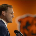 Ben Johnson in blauem Anzug und gelber Krawatte steht lächelnd da und hält ein NFL-Mikrofon in der Hand. Im Hintergrund ist eine orangefarbene Wand mit einem teilweise sichtbaren großen Bärenlogo zu sehen. Die Szene, die möglicherweise Ben Johnson bei einer Pressekonferenz zeigt, deutet auf eine sportbezogene Ankündigung hin. Diese Beschreibung wurde mit der FootballR KI automatisch generiert.