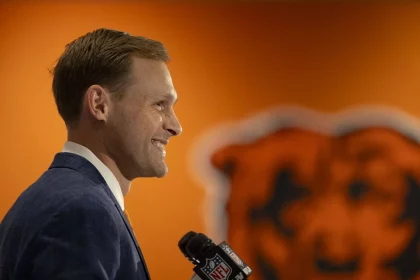 Ben Johnson in blauem Anzug und gelber Krawatte steht lächelnd da und hält ein NFL-Mikrofon in der Hand. Im Hintergrund ist eine orangefarbene Wand mit einem teilweise sichtbaren großen Bärenlogo zu sehen. Die Szene, die möglicherweise Ben Johnson bei einer Pressekonferenz zeigt, deutet auf eine sportbezogene Ankündigung hin. Diese Beschreibung wurde mit der FootballR KI automatisch generiert.