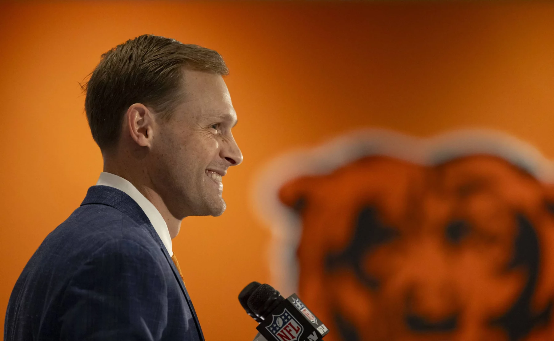 Ben Johnson in blauem Anzug und gelber Krawatte steht lächelnd da und hält ein NFL-Mikrofon in der Hand. Im Hintergrund ist eine orangefarbene Wand mit einem teilweise sichtbaren großen Bärenlogo zu sehen. Die Szene, die möglicherweise Ben Johnson bei einer Pressekonferenz zeigt, deutet auf eine sportbezogene Ankündigung hin. Diese Beschreibung wurde mit der FootballR KI automatisch generiert.