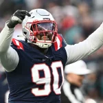 American-Football-Spieler Christian Barmore trägt ein dunkelblaues Trikot mit der Nummer 90 und einen Helm und reckt jubelnd die Arme in die Höhe. Im Hintergrund sind Zuschauer und ein Schiedsrichter zu sehen. Er trägt rot-weiße Streifen und das Logo eines Profiteams. Diese Beschreibung wurde mit der FootballR KI automatisch generiert.