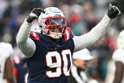 American-Football-Spieler Christian Barmore trägt ein dunkelblaues Trikot mit der Nummer 90 und einen Helm und reckt jubelnd die Arme in die Höhe. Im Hintergrund sind Zuschauer und ein Schiedsrichter zu sehen. Er trägt rot-weiße Streifen und das Logo eines Profiteams. Diese Beschreibung wurde mit der FootballR KI automatisch generiert.