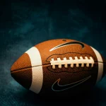 Eine Nahaufnahme eines American Footballs mit sichtbaren weißen Schnürsenkeln und Streifen sowie zwei Nike-Swoosh-Logos vor einem dunkelblauen, strukturierten Hintergrund. Der Football ist eine Anspielung auf die Vergünstigungen des NFL-Bonussystems, die sich die Top-Stars der Steelers auf dem Spielfeld verdienen. Diese Beschreibung wurde mit der FootballR KI automatisch generiert.