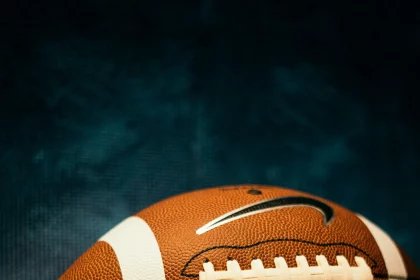 Eine Nahaufnahme eines American Footballs mit sichtbaren weißen Schnürsenkeln und Streifen sowie zwei Nike-Swoosh-Logos vor einem dunkelblauen, strukturierten Hintergrund. Der Football ist eine Anspielung auf die Vergünstigungen des NFL-Bonussystems, die sich die Top-Stars der Steelers auf dem Spielfeld verdienen. Diese Beschreibung wurde mit der FootballR KI automatisch generiert.