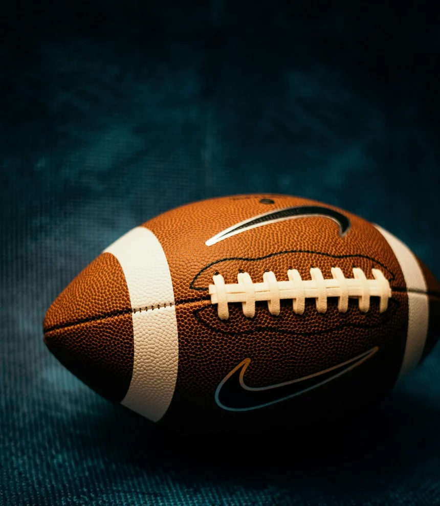Eine Nahaufnahme eines American Footballs mit sichtbaren weißen Schnürsenkeln und Streifen sowie zwei Nike-Swoosh-Logos vor einem dunkelblauen, strukturierten Hintergrund. Der Football ist eine Anspielung auf die Vergünstigungen des NFL-Bonussystems, die sich die Top-Stars der Steelers auf dem Spielfeld verdienen. Diese Beschreibung wurde mit der FootballR KI automatisch generiert.