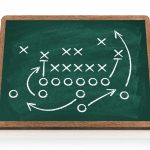Defensive Fronts - Eine Tafel zeigt ein Spieldiagramm im American Football. X und O stehen für gegnerische Spieler, Pfeile veranschaulichen Bewegung, Verteidigungslinien und Strategie. Blocken, Laufen, Deckung und Passrouten sind mit weißer Kreide auf einer grünen, holzgerahmten Tafel skizziert. Diese Beschreibung wurde mit der FootballR KI automatisch generiert.