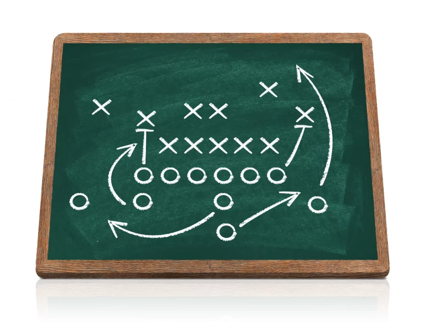 Defensive Fronts - Eine Tafel zeigt ein Spieldiagramm im American Football. X und O stehen für gegnerische Spieler, Pfeile veranschaulichen Bewegung, Verteidigungslinien und Strategie. Blocken, Laufen, Deckung und Passrouten sind mit weißer Kreide auf einer grünen, holzgerahmten Tafel skizziert. Diese Beschreibung wurde mit der FootballR KI automatisch generiert.