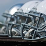 Eine Nahaufnahme von der Seite zeigt mehrere Footballhelme, meist weiß mit grauen Gesichtsmasken, die in einer Reihe auf einer Holzfläche aufgereiht sind. Der Hintergrund ist unscharf, was die Details und glänzenden Oberflächen der Helme hervorhebt. Diese Beschreibung wurde mit der FootballR KI automatisch generiert.