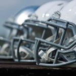 Eine Nahaufnahme von der Seite zeigt mehrere Footballhelme, meist weiß mit grauen Gesichtsmasken, die in einer Reihe auf einer Holzfläche aufgereiht sind. Der Hintergrund ist unscharf, was die Details und glänzenden Oberflächen der Helme hervorhebt. Diese Beschreibung wurde mit der FootballR KI automatisch generiert.