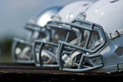 Eine Nahaufnahme von der Seite zeigt mehrere Footballhelme, meist weiß mit grauen Gesichtsmasken, die in einer Reihe auf einer Holzfläche aufgereiht sind. Der Hintergrund ist unscharf, was die Details und glänzenden Oberflächen der Helme hervorhebt. Diese Beschreibung wurde mit der FootballR KI automatisch generiert.