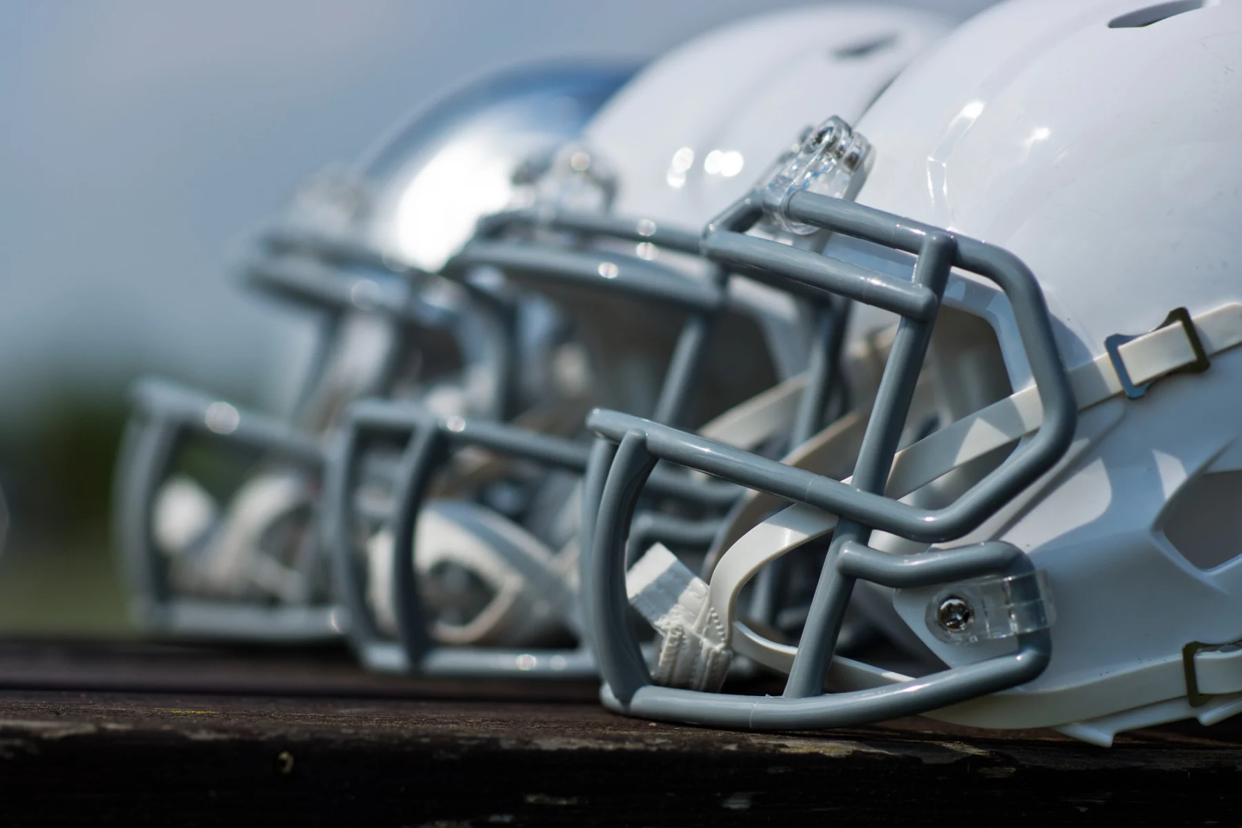 Eine Nahaufnahme von der Seite zeigt mehrere Footballhelme, meist weiß mit grauen Gesichtsmasken, die in einer Reihe auf einer Holzfläche aufgereiht sind. Der Hintergrund ist unscharf, was die Details und glänzenden Oberflächen der Helme hervorhebt. Diese Beschreibung wurde mit der FootballR KI automatisch generiert.