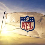 NFL Season 2025 - Eine weiße Flagge mit dem NFL-Schildlogo, auf dem ein Football und Sterne über den fettgedruckten roten Buchstaben „NFL“ zu sehen sind, weht im Wind vor einem hellen, goldenen Sonnenaufgangshimmel, in der Ferne sind am Horizont Berge zu sehen. Diese Beschreibung wurde mit der FootballR KI automatisch generiert.