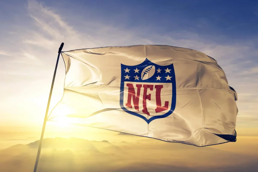 NFL Season 2025 - Eine weiße Flagge mit dem NFL-Schildlogo, auf dem ein Football und Sterne über den fettgedruckten roten Buchstaben „NFL“ zu sehen sind, weht im Wind vor einem hellen, goldenen Sonnenaufgangshimmel, in der Ferne sind am Horizont Berge zu sehen. Diese Beschreibung wurde mit der FootballR KI automatisch generiert.