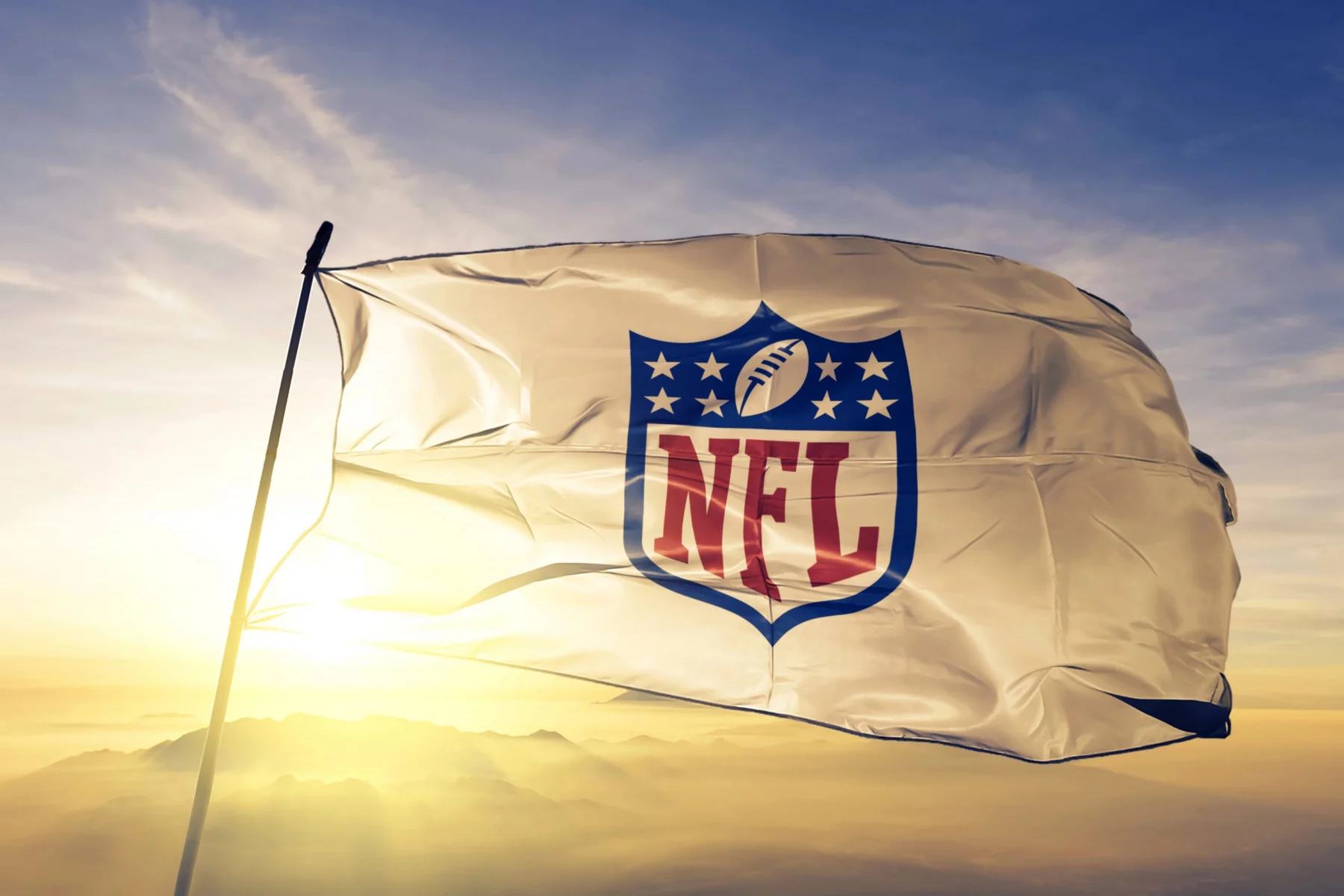 NFL Season 2025 - Eine weiße Flagge mit dem NFL-Schildlogo, auf dem ein Football und Sterne über den fettgedruckten roten Buchstaben „NFL“ zu sehen sind, weht im Wind vor einem hellen, goldenen Sonnenaufgangshimmel, in der Ferne sind am Horizont Berge zu sehen. Diese Beschreibung wurde mit der FootballR KI automatisch generiert.