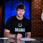 Maximilian Mang, Tight End der Indianapolis Colts, sitzt an einem Tisch und unterschreibt ein Dokument. Er trägt ein schwarzes T-Shirt mit einem weißen Hufeisen-Logo und der Aufschrift „MANG“. Hinter ihm steht eine Backsteinwand mit dem Colts-Logo und ein blau beleuchtetes Fenster. Diese Beschreibung wurde mit der FootballR KI automatisch generiert.