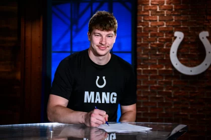 Maximilian Mang, Tight End der Indianapolis Colts, sitzt an einem Tisch und unterschreibt ein Dokument. Er trägt ein schwarzes T-Shirt mit einem weißen Hufeisen-Logo und der Aufschrift „MANG“. Hinter ihm steht eine Backsteinwand mit dem Colts-Logo und ein blau beleuchtetes Fenster. Diese Beschreibung wurde mit der FootballR KI automatisch generiert.