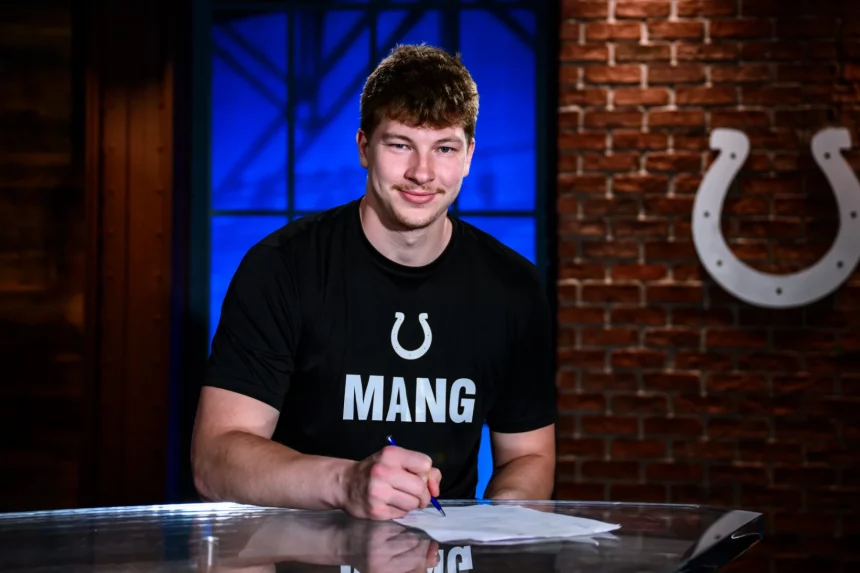 Maximilian Mang, Tight End der Indianapolis Colts, sitzt an einem Tisch und unterschreibt ein Dokument. Er trägt ein schwarzes T-Shirt mit einem weißen Hufeisen-Logo und der Aufschrift „MANG“. Hinter ihm steht eine Backsteinwand mit dem Colts-Logo und ein blau beleuchtetes Fenster. Diese Beschreibung wurde mit der FootballR KI automatisch generiert.