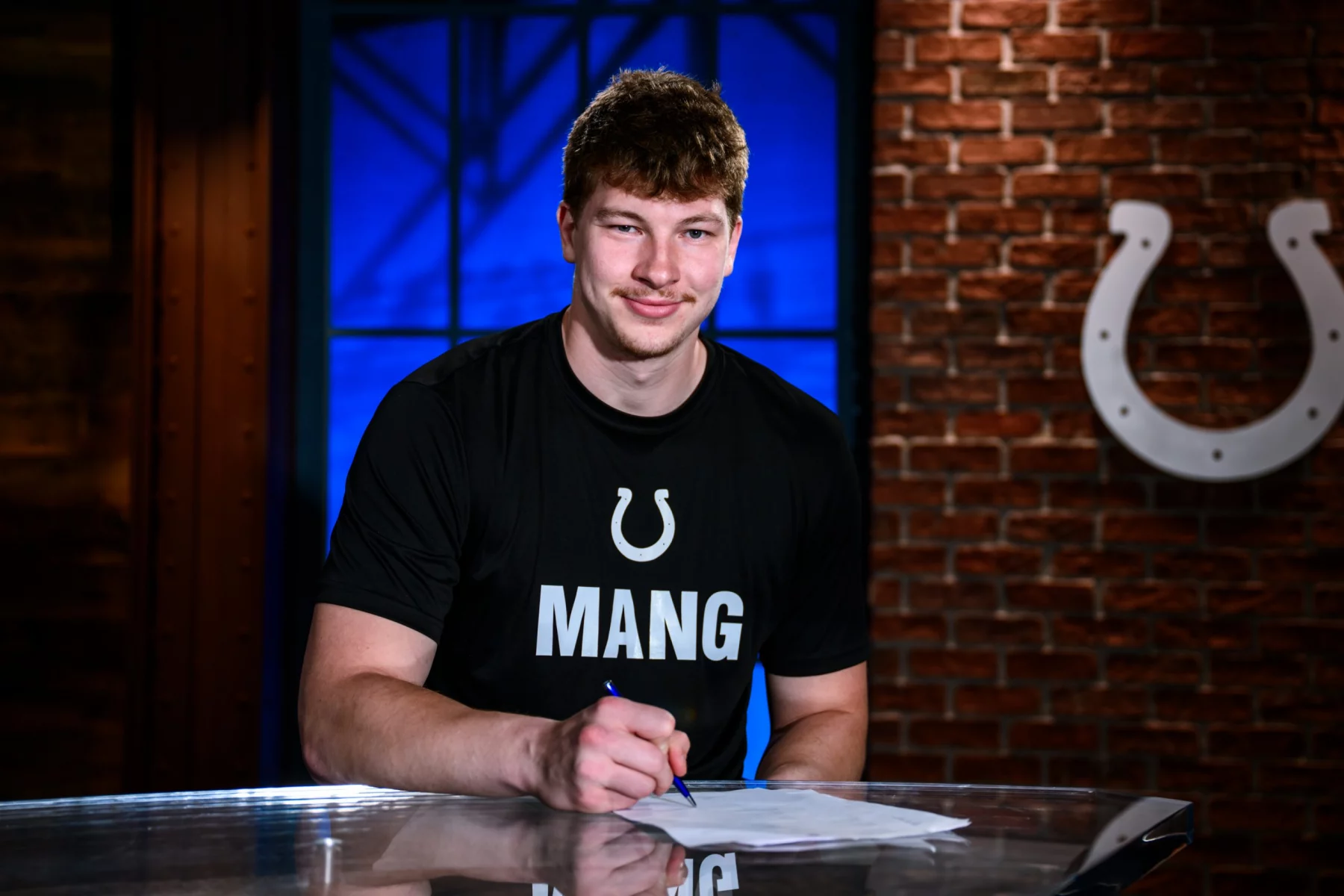 Maximilian Mang, Tight End der Indianapolis Colts, sitzt an einem Tisch und unterschreibt ein Dokument. Er trägt ein schwarzes T-Shirt mit einem weißen Hufeisen-Logo und der Aufschrift „MANG“. Hinter ihm steht eine Backsteinwand mit dem Colts-Logo und ein blau beleuchtetes Fenster. Diese Beschreibung wurde mit der FootballR KI automatisch generiert.