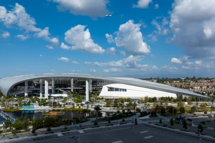 Flag Football - Eine Weitwinkelansicht des SoFi-Stadions unter blauem Himmel mit vereinzelten Wolken. Sein modernes, geschwungenes Dach dominiert die Szenerie. Hier wird Flag Football 2028 ausgetragen. In der Nähe befinden sich Grünflächen und Wasseranlagen, im Vordergrund ein leerer Parkplatz. Diese Beschreibung wurde mit der FootballR KI automatisch generiert.