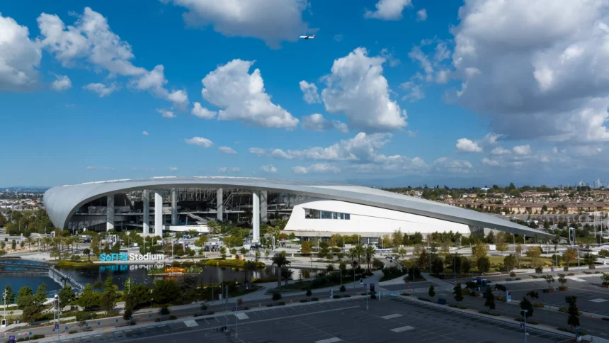 Flag Football - Eine Weitwinkelansicht des SoFi-Stadions unter blauem Himmel mit vereinzelten Wolken. Sein modernes, geschwungenes Dach dominiert die Szenerie. Hier wird Flag Football 2028 ausgetragen. In der Nähe befinden sich Grünflächen und Wasseranlagen, im Vordergrund ein leerer Parkplatz. Diese Beschreibung wurde mit der FootballR KI automatisch generiert.